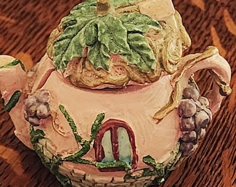 May include: A pink teapot shaped like a house with a green leafy roof and a small window. The teapot is decorated with vines and grapes.