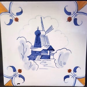 Könnte beinhalten: Eine handbemalte Keramikfliese in Blau und Weiß, die eine Windmühle in einer holländischen Landschaft darstellt. Die Fliese hat einen weißen Hintergrund mit blauen und orangen Akzenten.