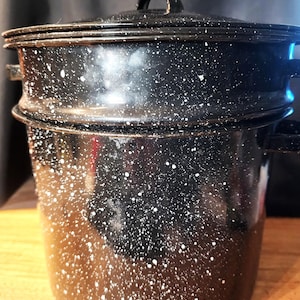 May include: A black enamel stockpot with a white speckled pattern, featuring a matching lid with a central handle and two side handles. The cylindrical pot has a smooth, glossy finish and a distinct rim where the lid rests securely.