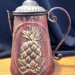 Puede incluir: Una tetera decorativa de metal con un acabado envejecido en tonos rojos, marrones y dorados. La tetera presenta un diseño de piña en relieve en la parte delantera, un asa curva y una tapa con un pequeño pomo. La tetera es decorativa.