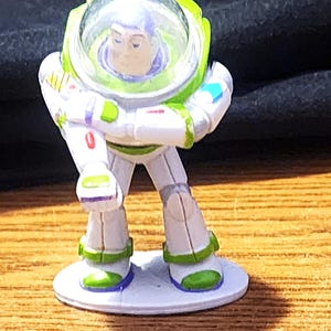 May include: A Buzz Lightyear figurine from the Toy Story franchise. The toy is primarily white with green and purple accents. It features a clear helmet and is standing on a small, round, white base. The figurine is posed with arms crossed.
