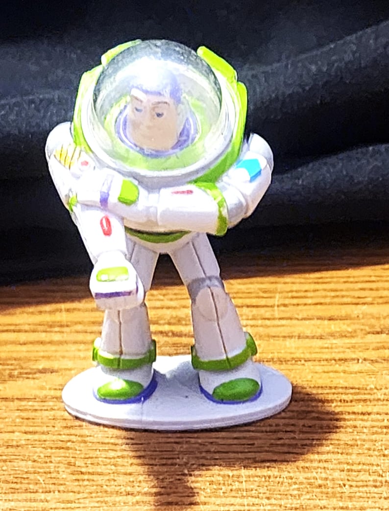 May include: A Buzz Lightyear figurine from the Toy Story franchise. The toy is primarily white with green and purple accents. It features a clear helmet and is standing on a small, round, white base. The figurine is posed with arms crossed.