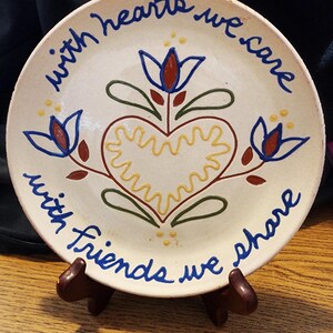 May include: A white ceramic plate with blue and yellow painted flowers and a heart. The plate has the text "with hearts we care with friends we share" written around the edge.