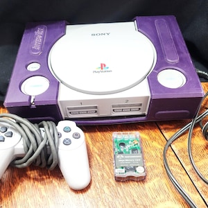 May include: A retro purple and white PlayStation console, accompanied by two white controllers and a memory card. The console features the PlayStation logo and the word "SONY". The controllers are linked to the console via grey cables.