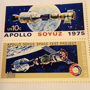Op de afbeelding: Twee postzegels van de Verenigde Staten ter herdenking van het Apollo-Sojoez-testproject. De bovenste postzegel heeft een blauwe achtergrond met een witte wereld en twee ruimtevaartuigen, een met het label "Apollo" en de andere met "Sojoez". De onderste postzegel heeft een zwarte achtergrond met een witte wereld en twee ruimtevaartuigen, een met het label "Apollo" en de andere met "Sojoez". Beide postzegels dragen de tekst "Apollo Soyuz Space Test Project" en "United States - 1975".