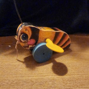 May include: A vintage wooden toy bee with a painted face, black and yellow striped body, and yellow wings. The toy has a blue wheel and a red antenna. The toy is sitting on a wooden surface.