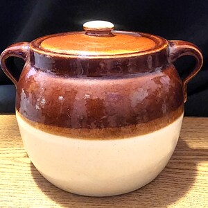 May include: A ceramic crock pot with a lid. The pot is two-toned, with a cream-colored base and a brown upper section. It has two handles and a brown lid with a small, round knob. The crock pot is designed for cooking and food storage.