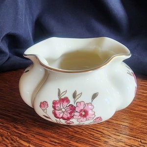 May include: A small, white ceramic creamer with a ruffled rim and a floral design in pink and gold. The creamer has a rounded shape and a slightly flared base.