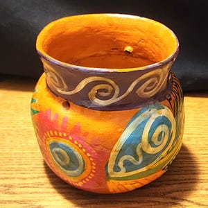 Könnte beinhalten: Ein kleiner, orangefarbener Tontopf mit einem violetten Rand und bemalt mit bunten Wirbeln und Blumen. Der Topf hat ein paar Löcher an der Seite.
