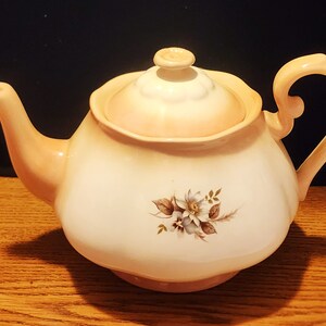 May include: A cream-coloured teapot with a floral design. The teapot has a lid and a handle. The floral design is in shades of brown and white.