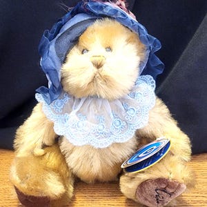 Peut inclure: Un ours en peluche brun portant un bonnet bleu avec une garniture en dentelle et un col en dentelle bleu. L'ours a des yeux en bouton brun et un nez brun. L'ours est assis sur une surface en bois.
