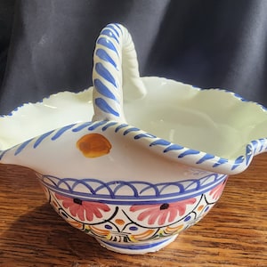 May include: A white ceramic bowl with a blue and white striped handle. The bowl has a scalloped edge and is decorated with blue, orange, yellow, and pink floral designs.