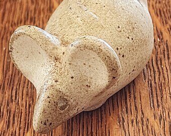 以下が含まれることがあります： 茶色と白の斑点のある釉薬をかけた、小さな陶器製のネズミの置物。ネズミは横向きに寝ていて、頭は右を向いている。