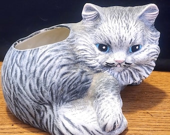 May include: A ceramic planter shaped like a gray and white cat with blue eyes. The cat is lying down with its head resting on its paws. The planter has a small opening on the top for planting.