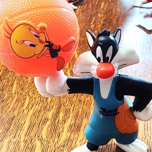 May include: A Sylvester the Cat figurine holding an orange basketball with a Tweety Bird graphic. Sylvester is wearing a blue and orange uniform with the text "Tune Squad". The figurine is on a wooden surface.