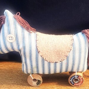May include: A stuffed horse toy made from blue and white striped fabric with a brown yarn mane and tail. It features a white button eye, a tan burlap saddle patch, and four wooden thread spools as wheels, with one spool labeled Coats & Clark.