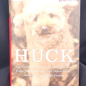 May include: A book titled HUCK by Janet Elder featuring a photograph of a light brown dog on the cover. The text reads: HUCK, The Remarkable True Story of How One Lost Puppy Taught a Family—and a Whole Town—About Hope and Happy Endings, JANET ELDER.