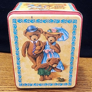 May include: A vintage tin box with a colorful illustration of three teddy bears. The bears are wearing clothes and the box has a red lid and a yellow base.