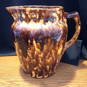 May include: A vintage brown and tan ceramic pitcher with a handle. The pitcher has a glossy finish and a mottled pattern of dark brown and tan. The pitcher is likely used for serving beverages and has a classic, rustic design.