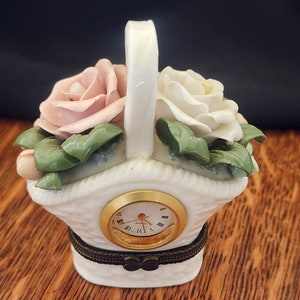 Puede incluir: Una cesta de porcelana blanca con asa, decorada con rosas rosas y blancas y hojas verdes. La cesta tiene una pequeña esfera de reloj con agujas doradas y números romanos.