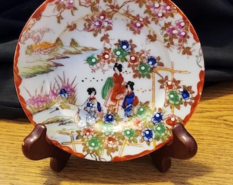 May include: A small, round, white porcelain plate with a red rim. The plate features a colorful Japanese-inspired design with three women in traditional clothing standing in a garden setting. The design includes flowers, trees, and a bridge.
