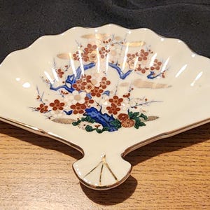 Puede incluir: Un plato de cerámica blanca en forma de abanico con un borde dorado. El plato presenta un diseño pintado a mano de un árbol azul y verde con flores rojas y blancas.