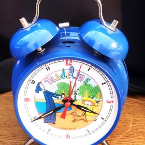 May include: A blue twin-bell alarm clock featuring a white face with the text Madeline. The clock face displays a colorful illustration of a character in a blue habit and a girl in a yellow hat with a dog. It has black hour and minute hands.