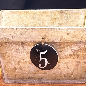 May include: A rectangular metal container with a weathered, textured finish. A black tag with the number "5" in white hangs from the container. The container is likely for storage or display.