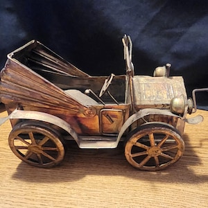 May include: A vintage metal model car with a bronze finish. The car has spoked wheels, a convertible top, and detailed headlights. The model is a decorative item, reminiscent of classic automobiles.