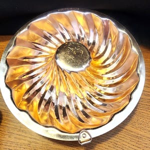 May include: A vintage, gold-toned bundt cake pan with a decorative spiral design. The pan has a central circular indentation and a silver-colored rim. A small metal clasp is visible on the edge. The pan is designed for baking cakes.