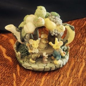 May include: A ceramic teapot figurine with a whimsical design. The teapot is decorated with a green, yellow, and brown floral motif and features four small mice figurines. The mice are dressed in different outfits and are positioned around the teapot.