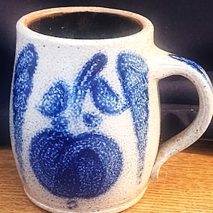 Pode incluir: Uma caneca cilíndrica de cerâmica com acabamento salpicado em branco sujo e superfície texturizada. A caneca apresenta motivos florais pintados à mão em azul cobalto e uma alça curva no lado direito. O interior da caneca é escuro e profundo.