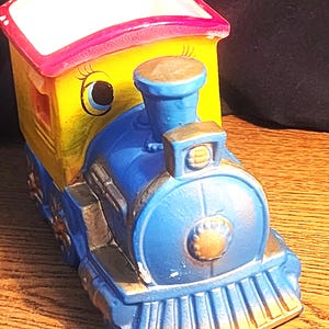 May include: A ceramic train-shaped planter featuring a blue locomotive front with a gold-toned cowcatcher and headlight. The cabin is yellow with a bright pink roof and includes a painted eye with eyelashes on the side of the engine structure.