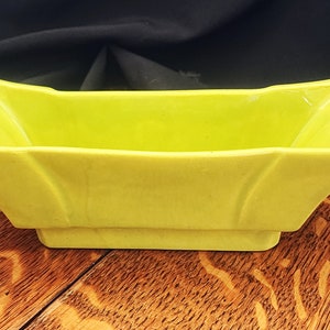 May include: A bright yellow ceramic planter with a rectangular shape and squared corners. The planter has a simple design with a slight inward curve at the top.