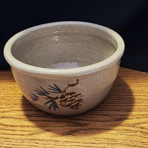 May include: A small, gray ceramic bowl with a brown pine cone design. The bowl is glazed and has a slightly textured surface.
