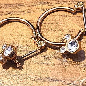 Puede incluir: Un par de pendientes de aro dorados. Cada pendiente presenta un dije con un cristal transparente facetado central y dos cristales m&aacute;s peque&ntilde;os, creando un dise&ntilde;o de tres cristales. Los pendientes est&aacute;n sobre una superficie de madera.