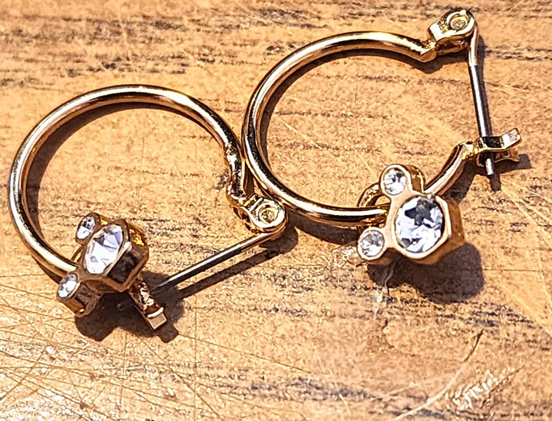 Puede incluir: Un par de pendientes de aro dorados. Cada pendiente presenta un dije con un cristal transparente facetado central y dos cristales m&aacute;s peque&ntilde;os, creando un dise&ntilde;o de tres cristales. Los pendientes est&aacute;n sobre una superficie de madera.