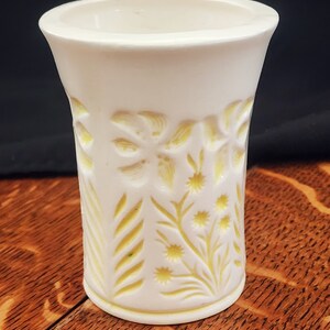 May include: White ceramic vase with a yellow floral design. The vase has a cylindrical shape and a smooth, glossy finish.