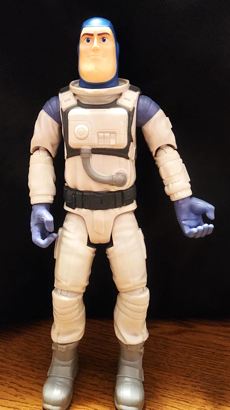 May include: A Buzz Lightyear action figure in a white and blue spacesuit. The figure has a blue helmet, gloves, and accents. The spacesuit has a white chest plate with a black belt and gray boots. The figure is standing upright.
