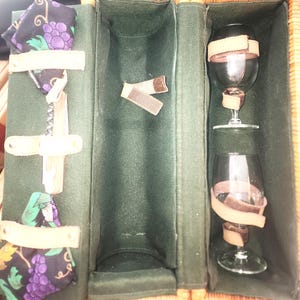 Puede incluir: Una cesta de picnic rectangular de mimbre con interior de tela verde oscuro. La cesta incluye dos copas de vino de cristal transparente sujetas con correas beige, un sacacorchos y dos servilletas con estampado de uvas moradas.