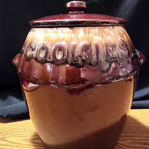 May include: A brown glazed ceramic cookie jar with a lid. The jar has the word "COOKIES" embossed on the front.