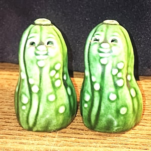 May include: A pair of green ceramic salt and pepper shakers shaped like pickles. Each shaker features a textured surface with raised white bumps, a rounded top with a small hole, and a molded face with eyes and a nose on the upper front section.
