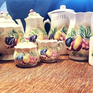 Op de afbeelding: Een vintage theeservies met een fruitmotief. De set bestaat uit een theepot, een roomkan, een suikerpot en vier kopjes. De onderdelen zijn versierd met een kleurrijk ontwerp van peren, pruimen en druiven.