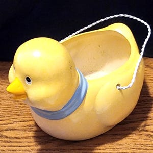May include: A yellow ceramic duck planter with a blue ribbon around its neck. The planter has a white rope handle and is designed to hold small plants or flowers.