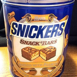 Könnte beinhalten: Eine blaue Blechdose mit Snickers-Snackriegeln. Die Dose hat ein gold-weißes Banner mit dem Wort "SNICKERS" in großen Buchstaben. Das Banner sagt auch "SNACK BARS" und "PACKED WITH PEANUTS". Die Dose enthält zwei 453g-Beutel.
