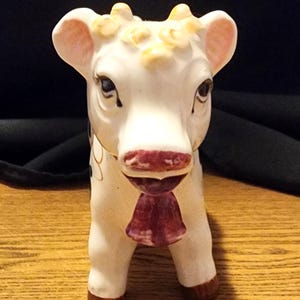 Może przedstawiać: Ceramiczna figurka krowy w kolorze białym z czarnymi plamami, żółtymi kręconymi włosami i różowym nosem. Krowa ma różową kokardkę wokół szyi.