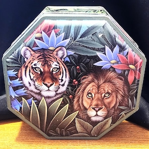 May include: An octagonal tin box featuring a detailed illustration of a tiger and a lion amidst lush foliage and colorful flowers. The box has a green border and a vintage aesthetic, perfect for storage or display.