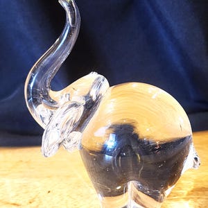 May include: A clear glass elephant figurine with a raised trunk. The elephant has a rounded body and is displayed on a wooden surface. The figurine is transparent, allowing light to pass through, and is a decorative item.