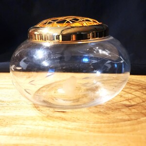 May include: A clear glass flower frog bowl featuring a delicate etched vine pattern on the exterior. The bowl is topped with a removable, polished gold-tone metal grid lid designed for arranging flowers, creating a functional and decorative piece.