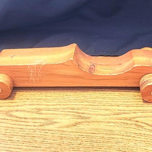 Könnte beinhalten: Ein handgefertigtes Holzspielzeugauto mit natürlicher Oberfläche. Das Auto hat eine geschwungene Karosserie, zwei Achsen und vier runde Räder. Das Spielzeugauto ist etwa 25 cm lang.